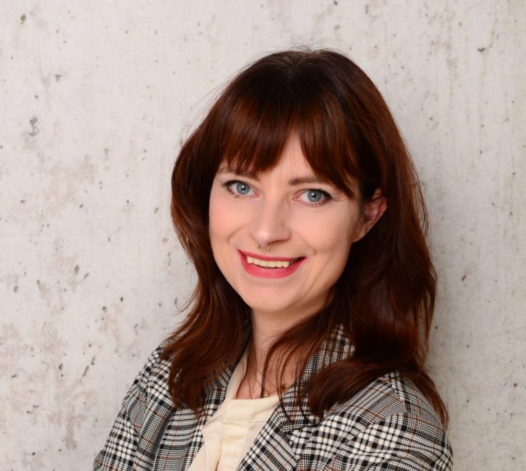 Bild von eine Frau mit schulter langem Haar, lächelt und trägt einen karierten Blazer Lächelnde Frau mit langen, braunen Haaren und kariertem Blazer vor hellem Hintergrund.